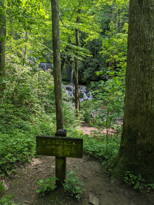 A sign for Virginia Hazzard Falls in a lush green forest, with a waterfall visible in the background.