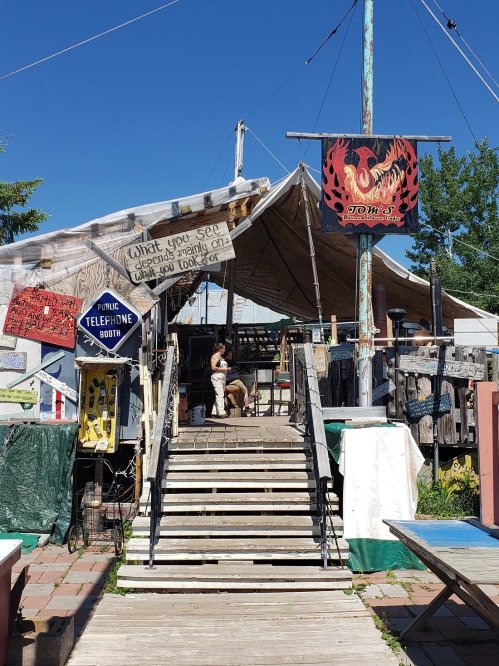 A rustic outdoor market with a large tent, wooden steps, and eclectic signs under a clear blue sky.