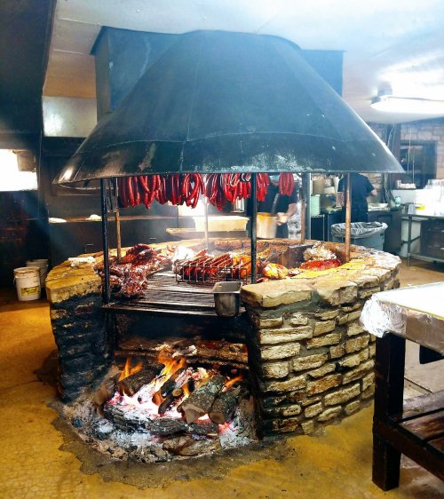 A rustic barbecue grill with meats and sausages hanging, surrounded by stone and a fire, in a cozy kitchen setting.
