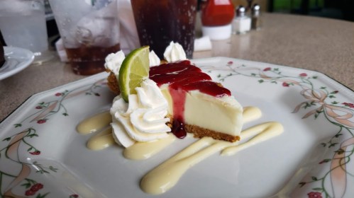 A slice of cheesecake topped with raspberry sauce, whipped cream, and a lime wedge, served on a decorative plate.