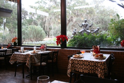 Cozy restaurant interior with tables set for dining, featuring a view of a garden and a sculpture outside.
