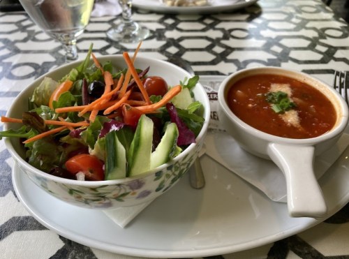 A bowl of fresh salad with mixed greens, tomatoes, and carrots, alongside a cup of tomato soup on a patterned table.