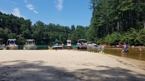 A sandy beach by a lake with boats docked and people enjoying the water and sun under a clear blue sky.