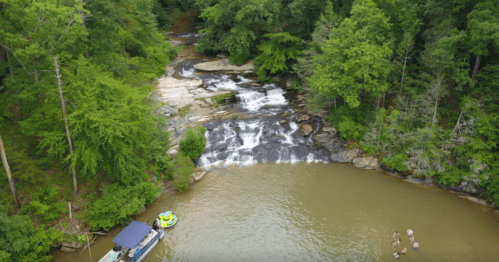 Aerial view of a serene waterfall flowing into a calm river, surrounded by lush green trees and a small boat nearby.