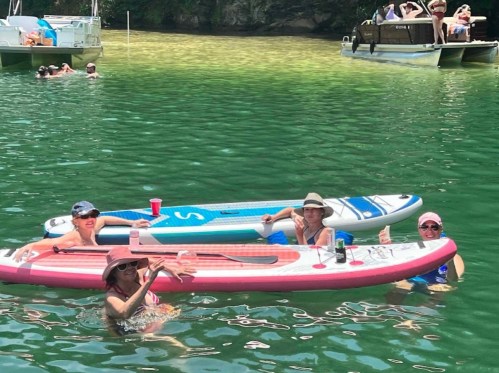 Three people on inflatable paddleboards in a green lake, enjoying drinks and giving thumbs up, with boats in the background.
