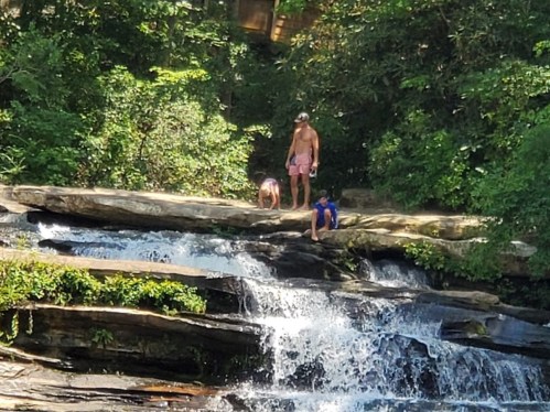 A man and two children explore a rocky area near a waterfall surrounded by lush greenery.