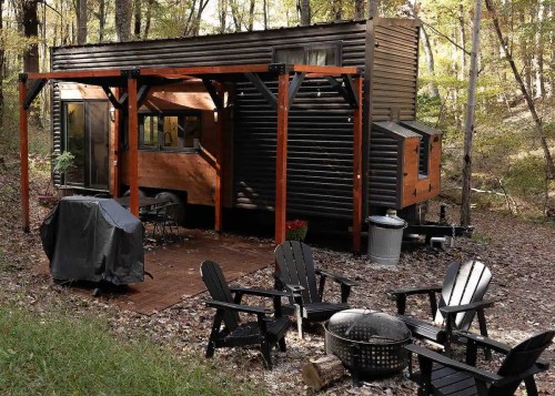 A modern tiny house in a wooded area, featuring a deck, outdoor seating, and a fire pit.