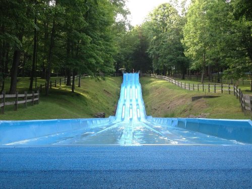 A blue water slide descends through a green park, surrounded by trees and a wooden fence.