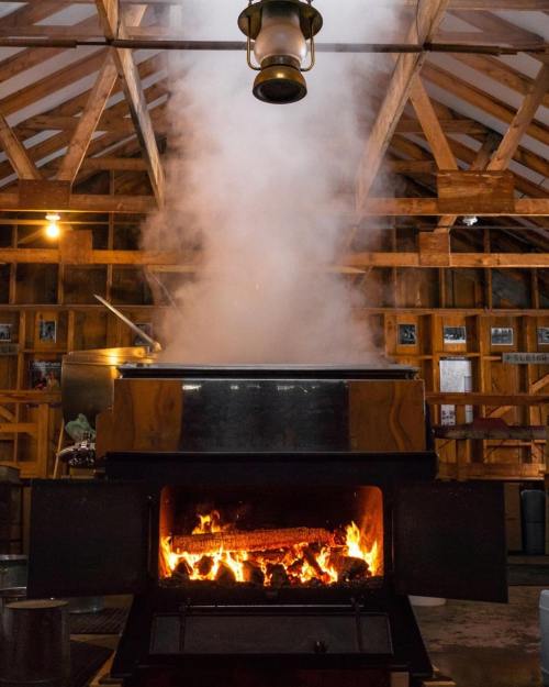 A wood-fired stove with flames and smoke rising in a rustic setting, surrounded by wooden beams and equipment.