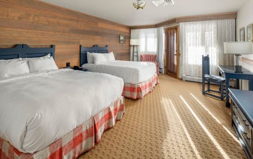 Cozy hotel room with two beds, wooden walls, and natural light from large windows. Warm, inviting decor.