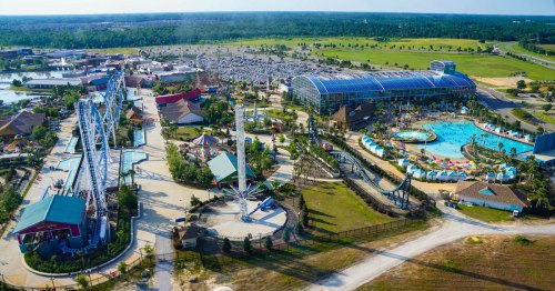 Aerial view of a large amusement park with rides, a water park, and extensive parking surrounded by greenery.