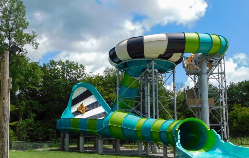 A colorful water slide with a spiral design, surrounded by green trees and a blue sky with clouds.