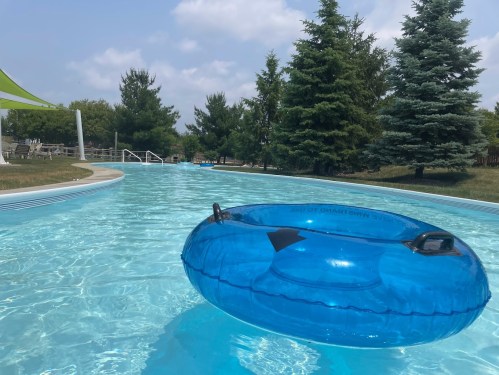 A blue inflatable tube floats in a clear swimming pool surrounded by green trees and a sunny sky.