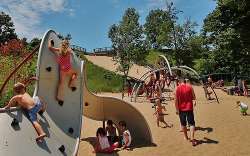 Children play on climbing structures in a sandy playground surrounded by greenery and trees on a sunny day.