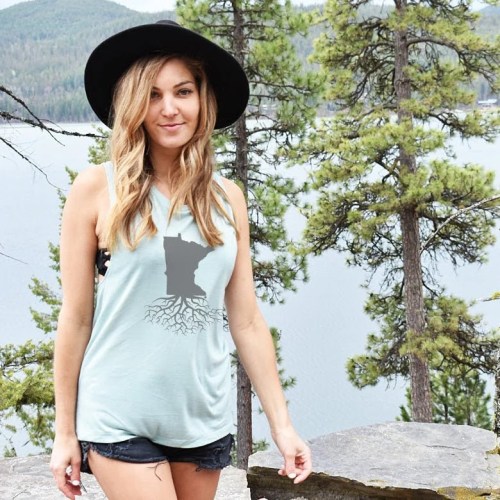 A woman in a tank top with a Minnesota graphic stands by a lake, surrounded by trees and mountains.