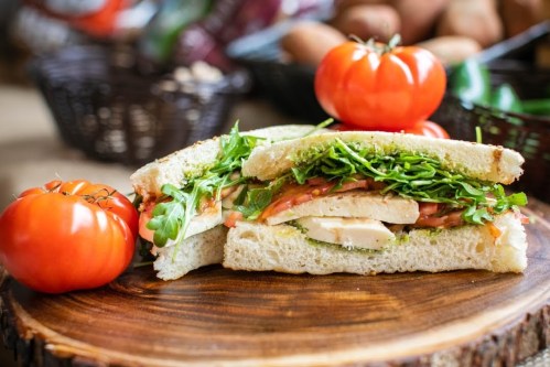 A fresh sandwich with chicken, tomatoes, and greens, served on a wooden board with tomatoes in the background.