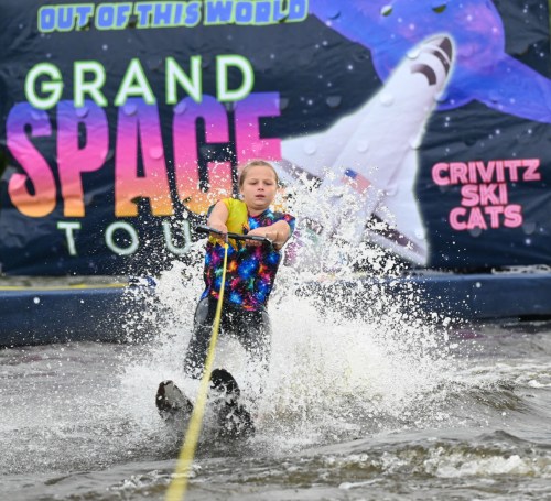 A young skier splashes through water while being towed, with a colorful "Grand Space Tour" banner in the background.