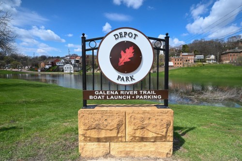Sign for Depot Park, featuring Galena River Trail, boat launch, and parking, with a scenic view of the river and blue sky.