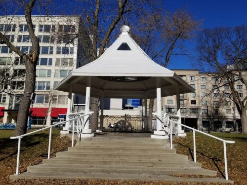 A white gazebo with a peaked roof, surrounded by trees and buildings, set against a clear blue sky.
