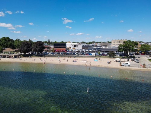 A sunny beach scene with people relaxing on the sand and a clear blue sky, alongside a waterfront and nearby buildings.