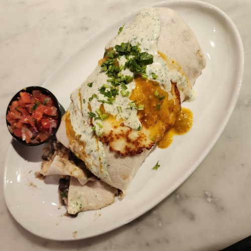 A large burrito topped with cheese and sauce, garnished with cilantro, served with a side of pico de gallo.