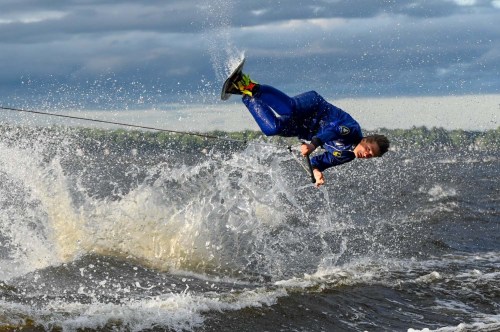A person in a blue wetsuit performs a trick while wakeboarding, splashing water against a cloudy sky backdrop.