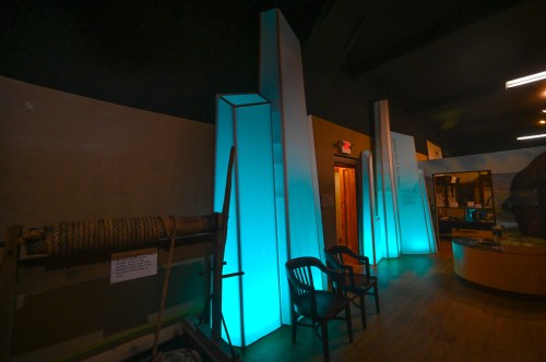 Interior of a museum featuring illuminated blue structures, wooden chairs, and a display area.