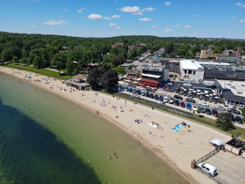Aerial view of a beach with people sunbathing, swimming, and a nearby town with shops and parked cars.