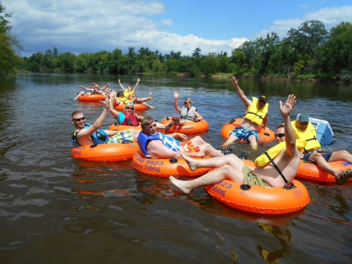 A group of people in orange inner tubes floating on a river, smiling and waving, surrounded by trees and blue skies.