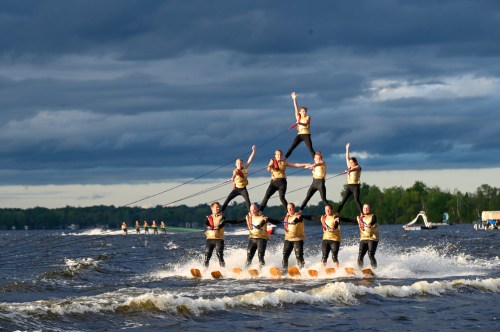 A pyramid of water skiers performing on a lake, with dark clouds in the background and boats in the distance.