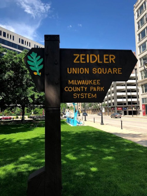 Signpost for Zeidler Union Square, part of the Milwaukee County Park System, with green grass and buildings in the background.