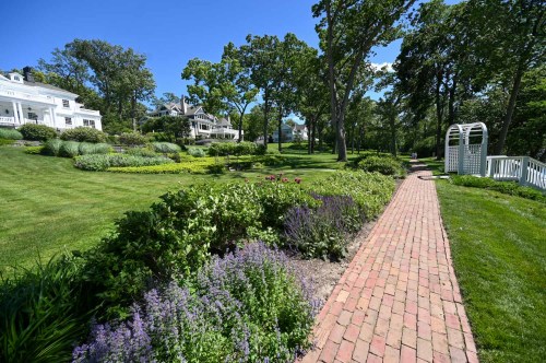 A scenic garden with a brick path, lush greenery, and blooming flowers, leading to a white gazebo and elegant houses.