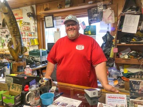 A smiling man in a red shirt stands behind a counter filled with fishing supplies and memorabilia in a rustic shop.
