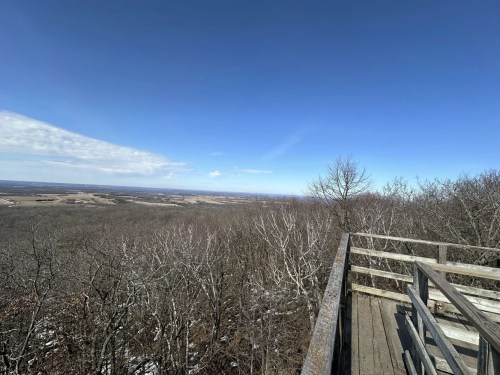 A wooden observation deck overlooks a vast landscape of trees and fields under a clear blue sky.