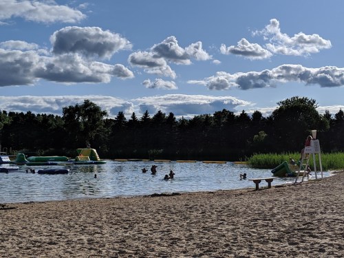A sandy beach with people swimming and playing in the water, a lifeguard on duty, and inflatable structures in the background.