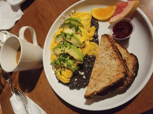 A plate with an omelette topped with avocado, black beans, toast, and fruit, alongside a cup of coffee.