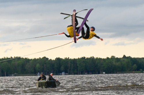 Two water skiers perform a jump over a lake, while a boat with two people watches nearby.