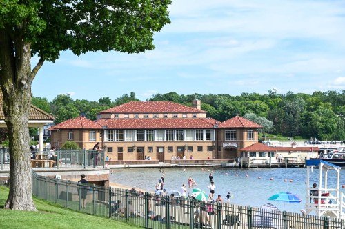 A lakeside scene featuring a large building, people enjoying the water, and green trees under a blue sky.