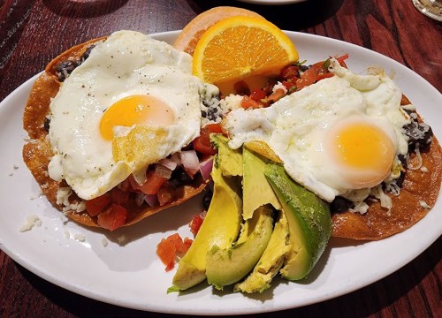 Two tostadas topped with eggs, black beans, diced tomatoes, onions, and avocado, served with orange slices.