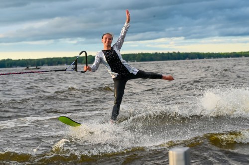 A person water skiing on a lake, smiling and balancing on one foot, with a scenic background of clouds and trees.