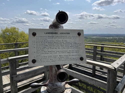 A viewpoint telescope with a landmark locator sign, overlooking a scenic landscape under a partly cloudy sky.