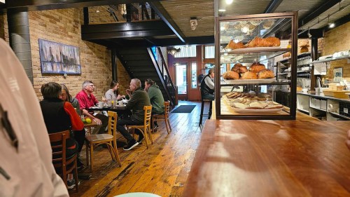 A cozy café interior with wooden floors, patrons enjoying meals, and a display case of pastries in the foreground.