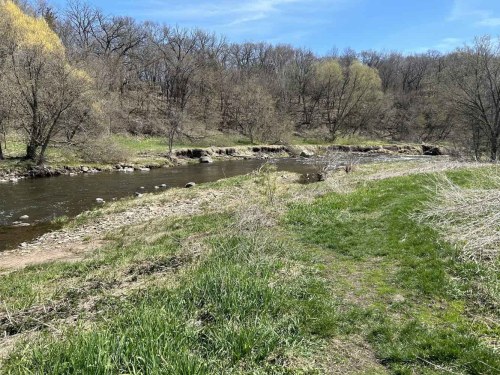 A serene riverbank scene with a flowing river, grassy areas, and trees in the background under a clear blue sky.