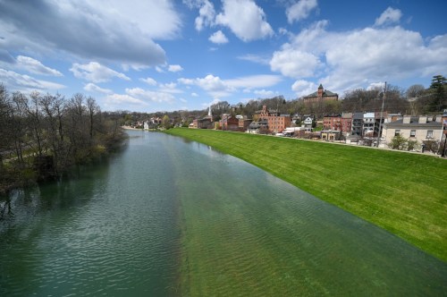 A serene river flows beside a grassy bank, with quaint buildings and trees under a partly cloudy sky.