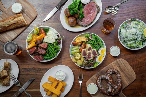 A table spread with various dishes: salads, meats, vegetables, and sides, alongside drinks and bread.