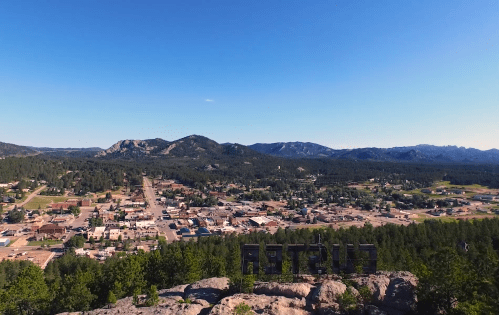 Aerial view of a small town surrounded by mountains and forests under a clear blue sky.