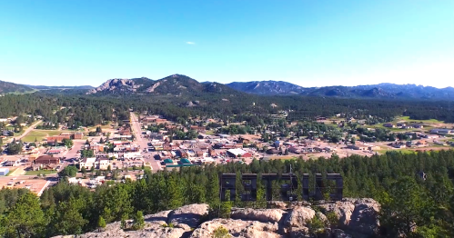 Aerial view of a small town surrounded by mountains and forests, with a sign on a rock in the foreground.