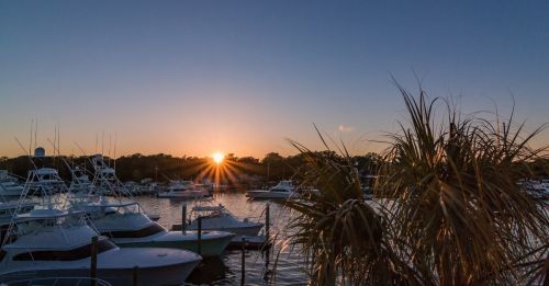 Sunset over a marina with boats and palm trees silhouetted against the colorful sky.