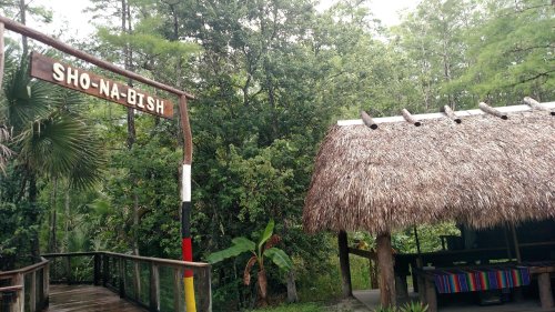 A wooden sign reading "Sho-Na-Bish" next to a thatched-roof structure in a lush, green forested area.