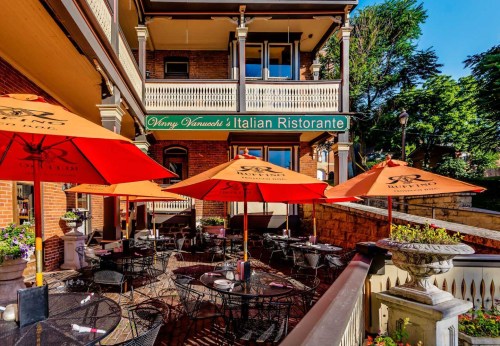 Outdoor dining area of an Italian restaurant with red umbrellas and tables, surrounded by greenery and historic architecture.
